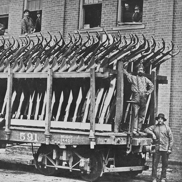 Fotografia de 1882 de três homens ao lado de um vagão da Deere & Co carregado de arados de aço a serem enviados e funcionários da fábrica olhando pela janela do prédio atrás