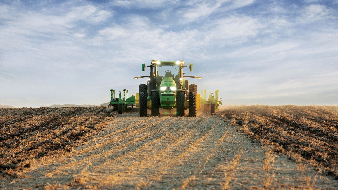 Trator John Deere com condução autónoma a puxar equipamento de lavoura num campo aberto.
