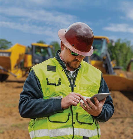 Trabalhador de construção com roupa de segurança verde olhando o celular na frente de uma carregadeira e um caminhão basculante.