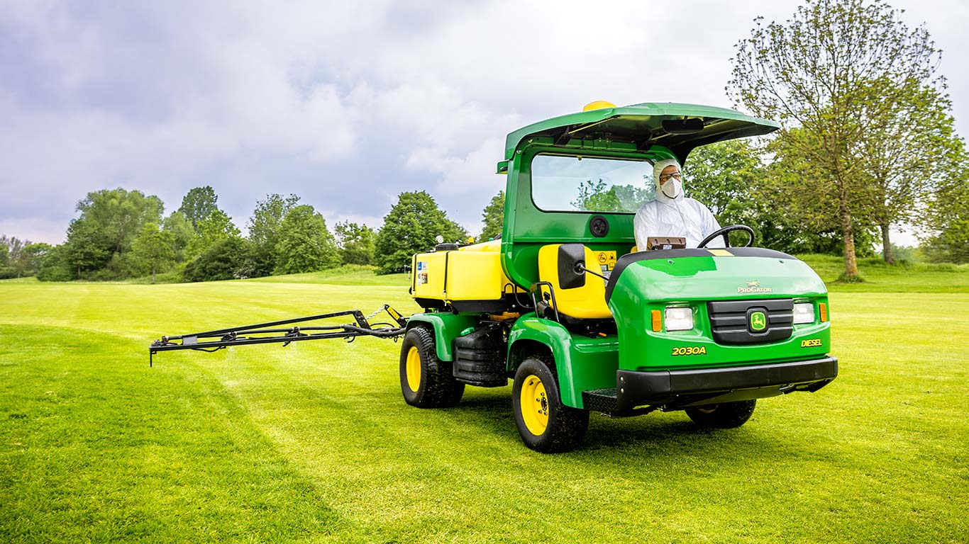 Um greenkeeper com vestuário protetor está a utilizar um pulverizador da John Deere no relvado de um campo de golfe.