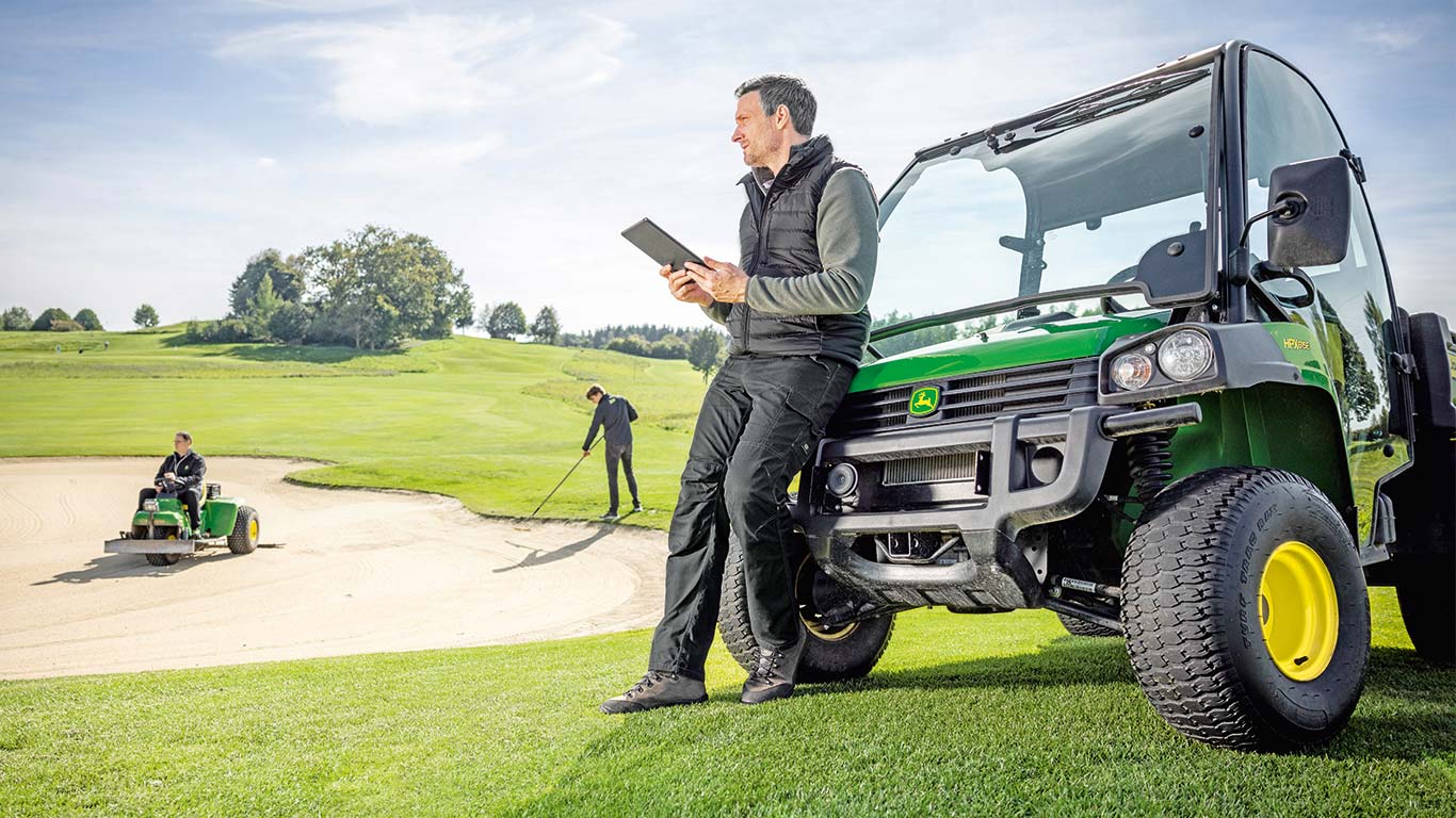 Um supervisor de greenkeepers está a verificar os dados no seu tablet enquanto 2 greenkeepers estão a trabalhar num campo de golfe ao fundo.