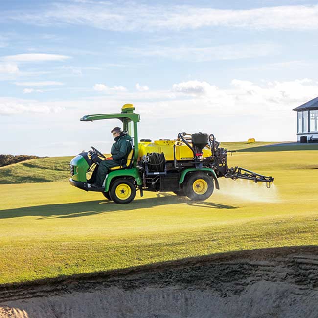 Um greenkeeper está a utilizar um pulverizador da John Deere numa área de relva