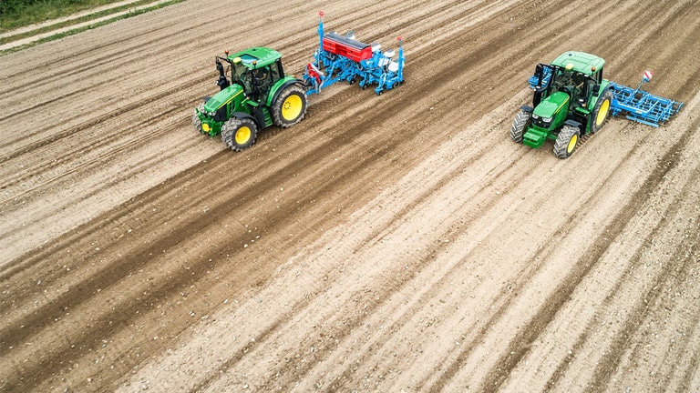 Dois tratores John Deere 6M num campo de terra. Um est&aacute; a arrastar um semeador Monsomen.  O outro est&aacute; a puxar um combinado de sementeiras Lemken