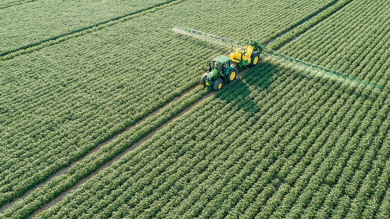 Um trator s&eacute;rie John Deere 6M a arrastar um pulverizador rebocado M700 num campo de batatas
