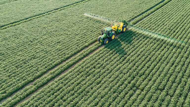 Um trator s&eacute;rie John Deere 6M a arrastar um pulverizador rebocado M700 num campo de batatas